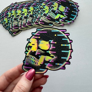 May include: A stack of colorful skull stickers with a glittery, pixelated design. The skull is outlined in black and has a yellow eye with a blue and purple glitch effect.