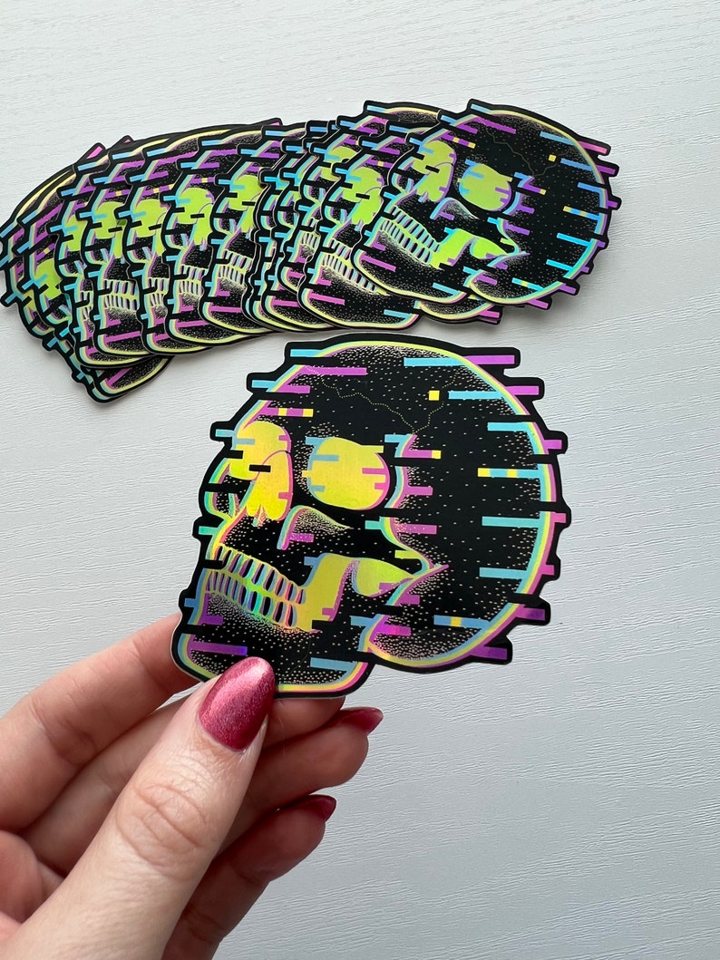 May include: A stack of colorful skull stickers with a glittery, pixelated design. The skull is outlined in black and has a yellow eye with a blue and purple glitch effect.