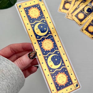 May include: A celestial-themed bookmark featuring a sun, moon, and stars design. The bookmark has a gold border with a navy blue background. The sun and moon have a face. The bookmark is rectangular and has a glossy finish.