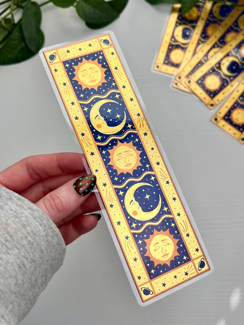 May include: A celestial-themed bookmark featuring a sun, moon, and stars design. The bookmark has a gold border with a navy blue background. The sun and moon have a face. The bookmark is rectangular and has a glossy finish.