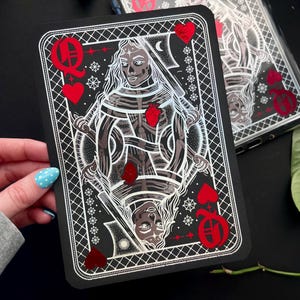 May include: A black playing card featuring a skeletal Queen of Hearts design with red heart accents. The card has a white border and intricate details. A phone case with a similar design is visible in the background.