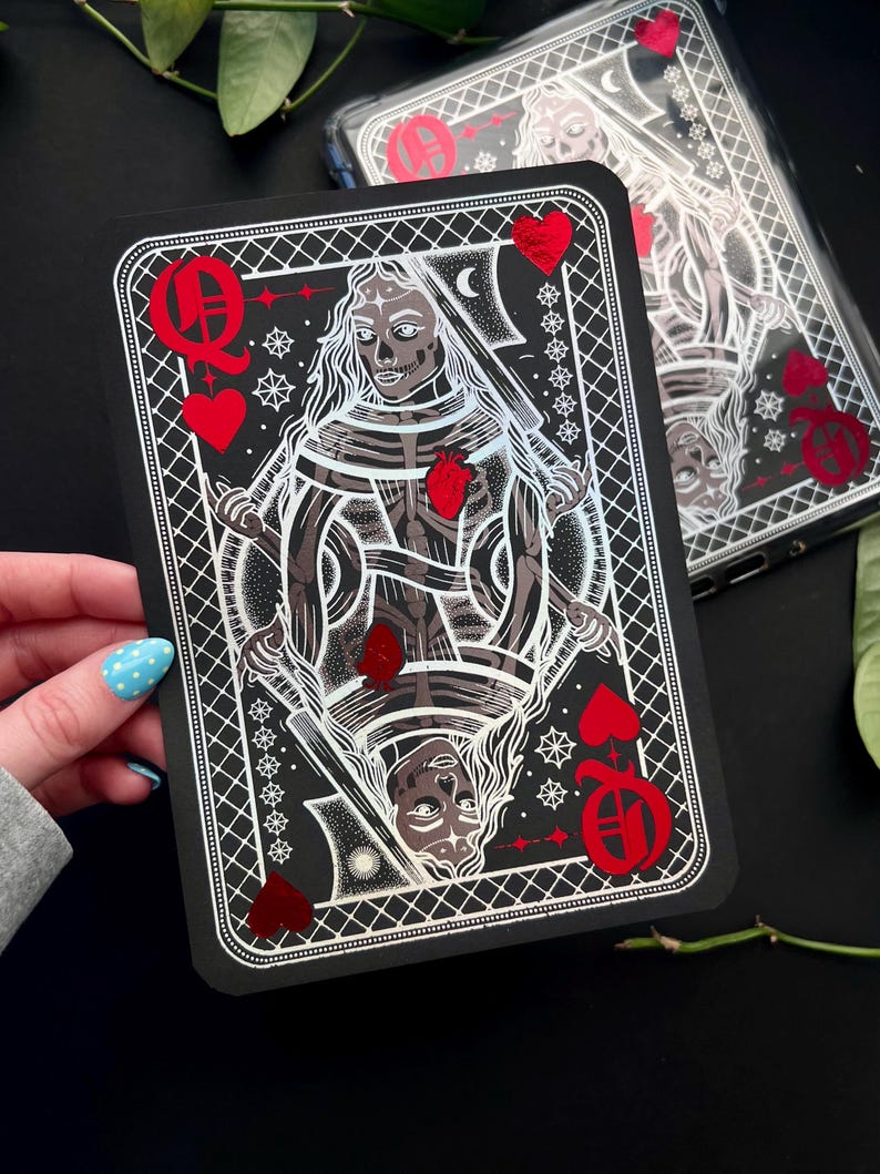 May include: A black playing card with a skeletal Queen of Hearts design and red heart details. The card has a white border and detailed artwork. A phone case with a similar design is visible in the background.