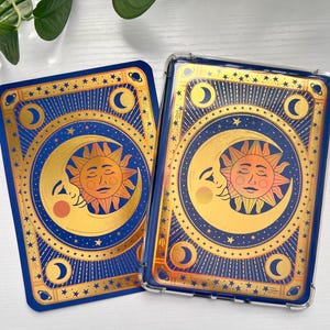 May include: Two rectangular cases with a celestial design. The cases feature a blue background with gold accents, including a crescent moon, sun, and stars. One case is encased in a clear protective cover.