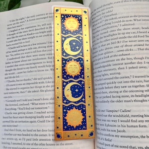 May include: A rectangular bookmark with a celestial design featuring gold suns, moons, and stars on a navy blue background. The bookmark is placed on top of an open book with text.