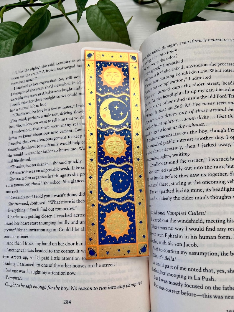 May include: A rectangular bookmark with a celestial design featuring gold suns, moons, and stars on a navy blue background. The bookmark is placed on top of an open book with text.