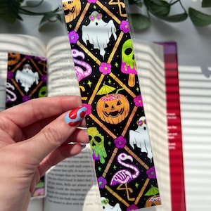 May include: A rectangular bookmark featuring a black background with a repeating diamond pattern in gold. The design includes white ghosts wearing heart-shaped glasses, orange jack-o'-lanterns with cocktail umbrellas, green melting skulls, and pink flamingo skeletons.