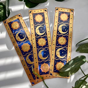 May include: Three celestial-themed bookmarks. Each bookmark features a gold sun and crescent moon design on a navy blue background, framed by a gold border with stars. The bookmarks are rectangular and appear to be made of a clear, glossy material.