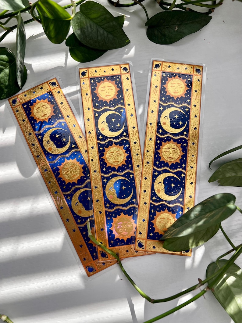 May include: Three celestial-themed bookmarks. Each bookmark features a gold sun and crescent moon design on a navy blue background, framed by a gold border with stars. The bookmarks are rectangular and appear to be made of a clear, glossy material.