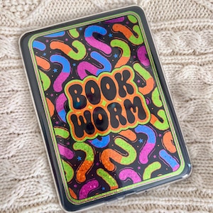 May include: A rectangular e-reader case featuring a black background with a vibrant, glittery pattern of colorful, curved worm shapes in neon green, orange, pink, and blue. The text BOOK WORM is centered in bold, orange, bubble-style letters.