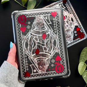 May include: Clear phone case with a Queen of Hearts playing card design. The case has a black background with a skeletal figure holding a sword, red hearts, and decorative elements. Another case is visible in the background.