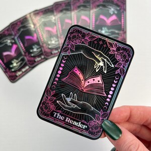 May include: A holographic tarot card with a black background and pink and purple accents. The card depicts two hands holding an open book with hearts on the pages. The text "The Reader" is printed at the bottom of the card.