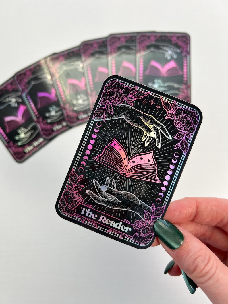 May include: A holographic tarot card with a black background and pink and purple accents. The card depicts two hands holding an open book with hearts on the pages. The text "The Reader" is printed at the bottom of the card.
