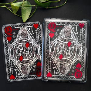 May include: Two playing cards featuring a Queen of Hearts design with a skeletal figure, red hearts, and intricate white detailing on a black background. One card is in a clear protective case.