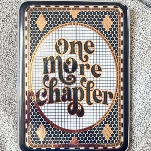 May include: A rectangular phone case with a black border and a decorative design. The design features the words "One More Chapter" in a vintage font, surrounded by a gold and black mosaic pattern. The background is white with a grid pattern.