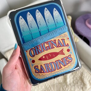 May include: A clear phone case with a design of a blue and yellow sardine can. The can has the text "ORIGINAL SARDINES" and a red fish illustration.