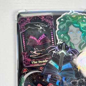 May include: A collection of holographic stickers featuring a tarot card with the text "The Reader", a sword in a book, a mushroom patch, a crescent moon, and a Medusa with green hair.