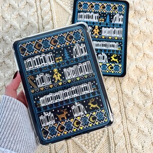 May include: A black and blue phone case with a wintery design featuring a bookshelf filled with books, a gingerbread man, a Christmas tree, and reindeer. The design is reminiscent of a knitted jumper.