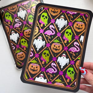 May include: A rectangular phone case and a matching print featuring a black background with a repeating lattice pattern. The design includes white ghosts with pink heart sunglasses, neon green melting skull popsicles, pink flamingo skeletons, and orange jack-o'-lanterns with green cocktail umbrellas.