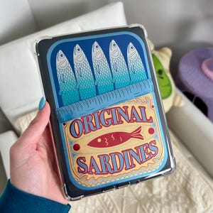 May include: A clear plastic case with a design of a blue and yellow sardine can. The can has five blue sardines and the text "ORIGINAL SARDINES" in red and yellow.