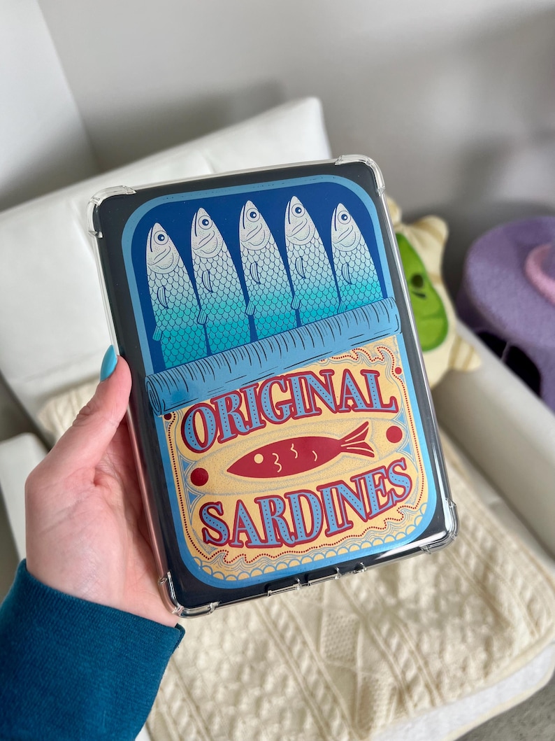 May include: A clear plastic case with a design of a blue and yellow sardine can. The can has five blue sardines and the text "ORIGINAL SARDINES" in red and yellow.