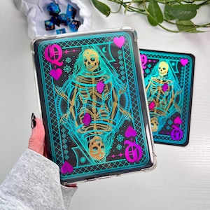Puede incluir: Una funda transparente con un diseño de carta de la Reina de Corazones, con una ilustración de esqueleto. La carta tiene un fondo negro con detalles turquesa y dorados, y símbolos de corazón rosa. Se ve otra funda con el mismo diseño.