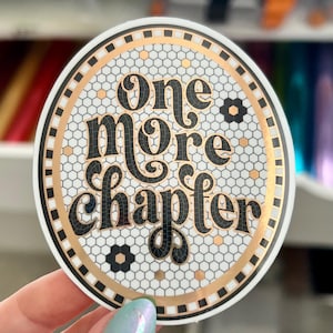 Puede incluir: Una pegatina redonda con el texto "One More Chapter" en negro. La pegatina tiene un fondo blanco con un patrón de panal y detalles dorados. El borde es negro y dorado.