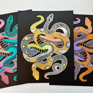 May include: Three black art prints with stylized snake illustrations. The snakes are in teal, gold, and purple, with celestial and geometric patterns. The artwork has a holographic, iridescent finish. The prints are suitable for framing.