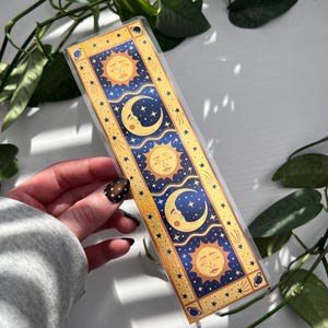 May include: A celestial-themed bookmark featuring sun and moon designs in gold, blue, and yellow. The bookmark has a rectangular shape with a clear plastic overlay. The design includes stars and celestial bodies, perfect for book lovers.