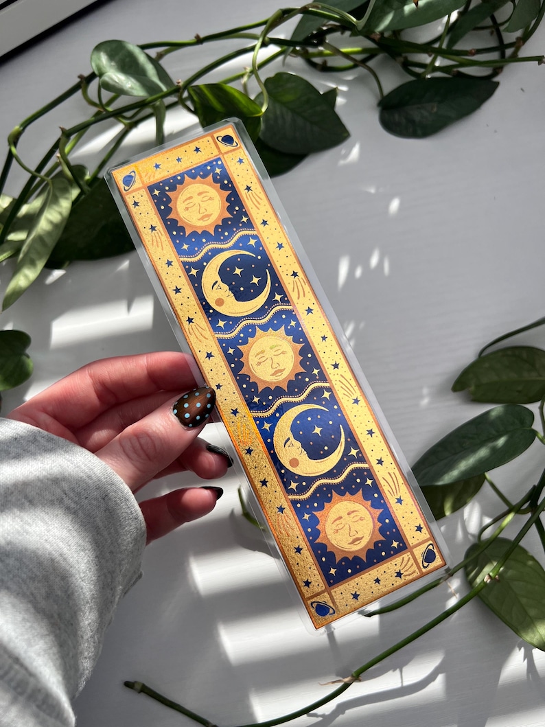 May include: A celestial-themed bookmark featuring sun and moon designs in gold, blue, and yellow. The bookmark has a rectangular shape with a clear plastic overlay. The design includes stars and celestial bodies, perfect for book lovers.