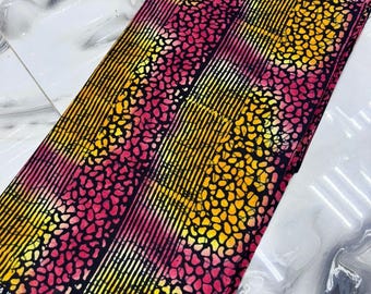 Op de afbeelding: Een gevouwen stuk stof met een kleurrijk patroon. Het ontwerp bevat verticale strepen afgewisseld met gespikkelde patronen in roze, geel en zwart. De stof heeft een label met tekst en een knoop.