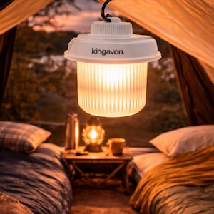 May include: A white camping lantern, branded "kingavon", hangs inside a tent. The lantern has a ribbed, cylindrical light diffuser. The tent's interior is illuminated with warm light, with beds and a small table visible in the background.