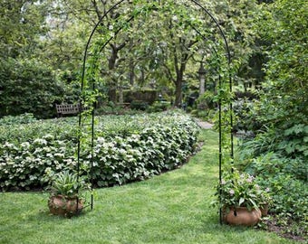 Op de afbeelding: Een zwarte metalen tuinboog met klimplanten, geplaatst in een weelderige groene tuin. Twee terracotta potten met planten staan aan de voet van de boog. Een pad leidt door de boog, omringd door groen.