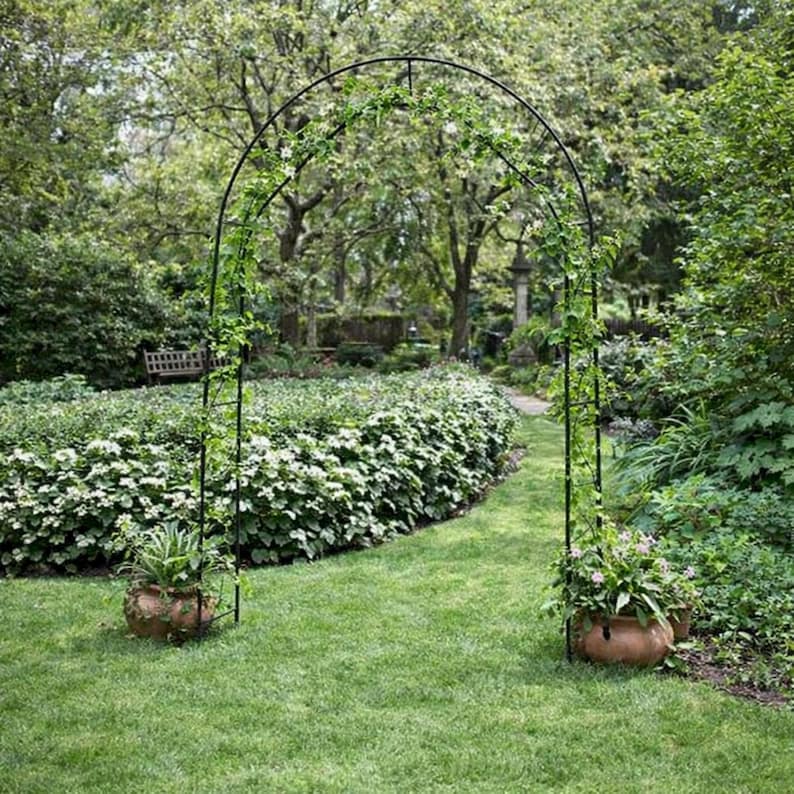 May include: A black metal garden archway with climbing plants, set in a lush green garden. Two terracotta pots with plants are at the base of the arch. A pathway leads through the arch, surrounded by greenery.