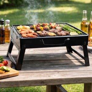 May include: A black portable charcoal grill with a variety of grilled foods, including shrimp, vegetables, and steak. The grill is set up on a wooden table outdoors, with bottles and beer visible in the background. The grill is approximately 30 cm wide.