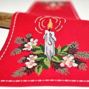 May include: A red Christmas table runner with an embroidered design. It features a white candle with an orange flame, surrounded by green foliage and white flowers. The runner has a white stitched border.