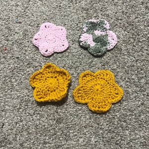 May include: Four crocheted flower appliques arranged in a square. One flower is solid light pink, one is a variegated mix of pink and dark green, and two are solid mustard yellow. Each flower features five rounded petals and a textured yarn surface.