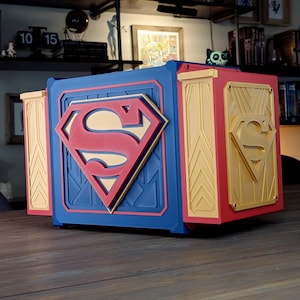 Peut inclure: Une boîte multicolore sur le thème de Superman avec un grand logo Superman sur le devant. La boîte est principalement bleue, rouge et dorée, avec le logo Superman bien en évidence. La boîte a un design géométrique.