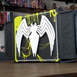 May include: A black and yellow decorative box featuring a white spider emblem. The box has a hinged design with a spiderweb pattern on the sides and a Venom-themed graphic. The box is displayed on a wooden surface.