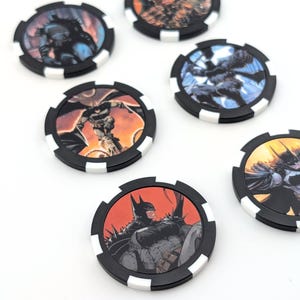 May include: Six circular poker-style chips featuring various graphic illustrations of Batman. Each chip has a black border with white rectangular accents along the edge. The center of each chip displays a different comic book style image of the character.