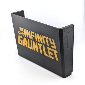 May include: Black and gold wall-mounted display featuring the text "THE INFINITY GAUNTLET". The display has a geometric design on the right side and is angled, with a white background.