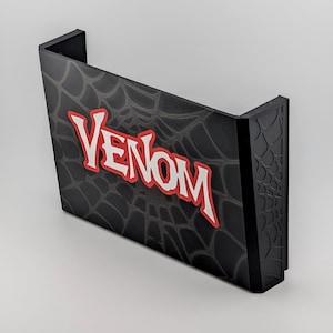 May include: A black, angular wall shelf featuring the word "Venom" in red and white lettering. The shelf has a spiderweb design on the front and a textured pattern on the side.