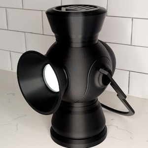 May include: A black lantern with a unique design, featuring a round body, a flared opening, and a handle. The lantern has a circular base and a top with a slotted design. The lantern is sitting on a white surface.