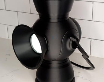 May include: A black lantern with a unique design, featuring a round body, a flared opening, and a handle. The lantern has a circular base and a top with a slotted design. The lantern is sitting on a white surface.