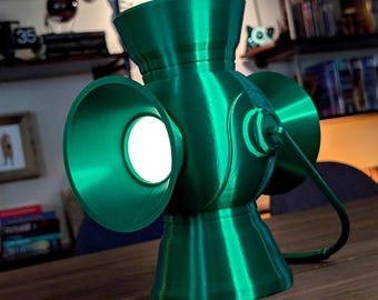 Peut inclure: Une lampe en forme de lanterne vert métallique avec un corps cylindrique lisse et trois ouvertures circulaires évasées. L'ouverture centrale émet une lumière blanche vive et un fin cordon d'alimentation noir sort du côté de la structure.