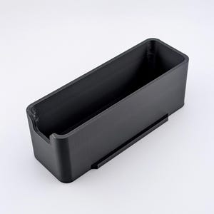 May include: A black rectangular container with a recessed area on one end and a small ledge on the side. The container is made of a solid, matte black material and sits on a white surface.