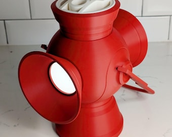 May include: A red lantern-shaped object with a white top and three circular openings. The lantern has a handle and a rounded base. The object is made of a smooth material and sits on a white surface.