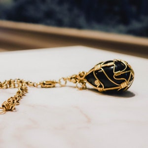 May include: A black teardrop-shaped pendant with gold wire detailing, suspended from a gold chain. The jewellery is presented on a white marble surface. The background is blurred, with a hint of a window.