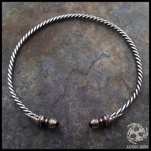 Puede incluir: Un collar gargantilla de alambre plateado trenzado con dos extremos decorativos que presentan una piedra roja oscura y diseños intrincados de plata.