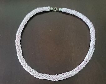 May include: A braided necklace composed of small, light-colored beads. The necklace features a delicate pattern with hints of pink and white, and is secured with a small clasp. The jewelry is displayed on a dark brown surface.