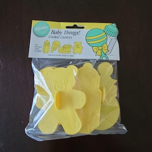 Puede incluir: Un juego de cuatro cortadores de galletas de plástico amarillo con forma de oso de peluche, biberón, sonajero y cochecito de bebé. El paquete dice "Baby Things! Cookie Cutters" y "Set Contains These 4 Cutters."